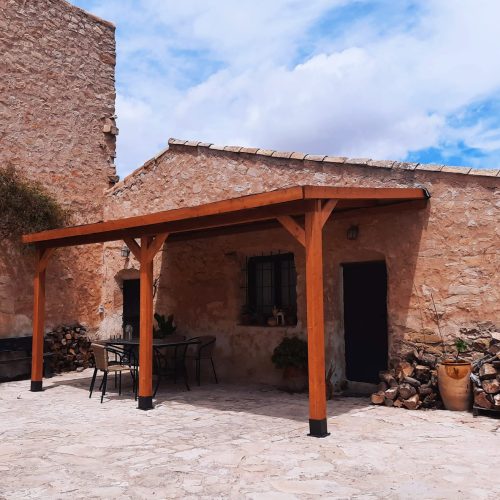 Pérgola de madera laminada adosada a pared con techo de madera y tela asfáltica, ideal para terrazas y patios. Estructura robusta en madera laminada tratada para exterior. Techo inclinado para una correcta evacuación del agua y mayor protección frente a lluvia y sol. Incluye anclajes metálicos al suelo para una instalación segura y estable. Perfecta para ampliar zonas de sombra, cubrir espacios exteriores y mejorar el entorno del hogar con un diseño premium, resistente y de bajo mantenimiento. Fabricada en España.